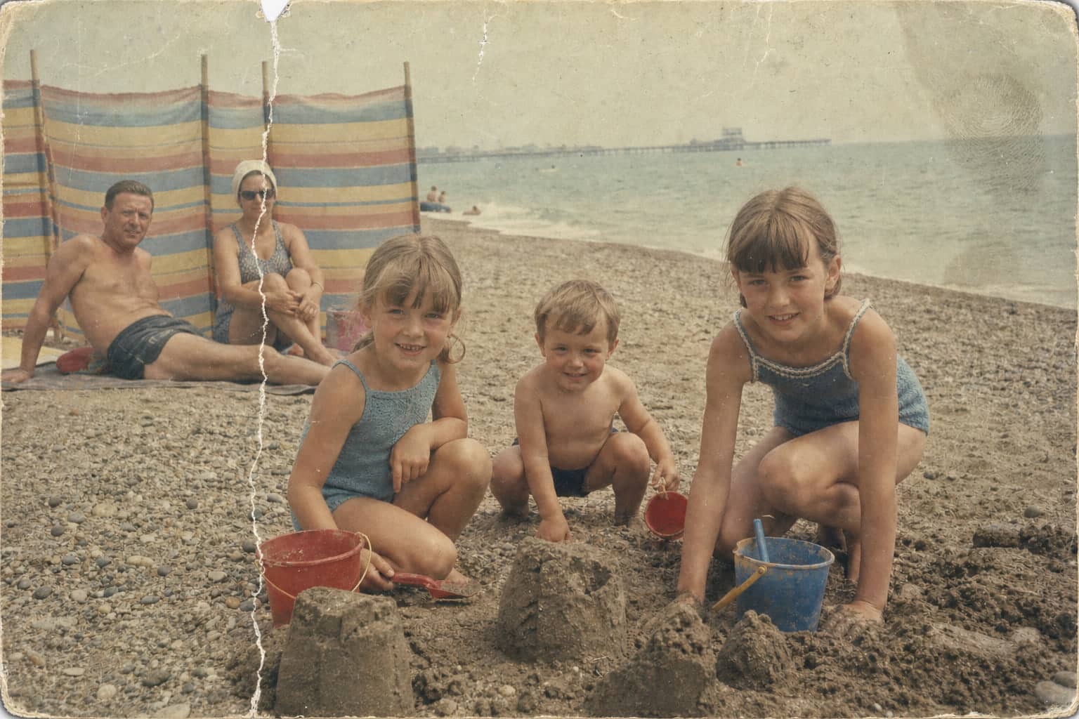 Faded, creased 1965 beach photo before restoration: three children by a sandcastle.