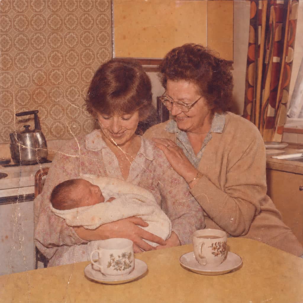 Creased, scuffed 1978 kitchen photo with an orange cast: a mother holding her newborn, her own mother beside her, before restoration.